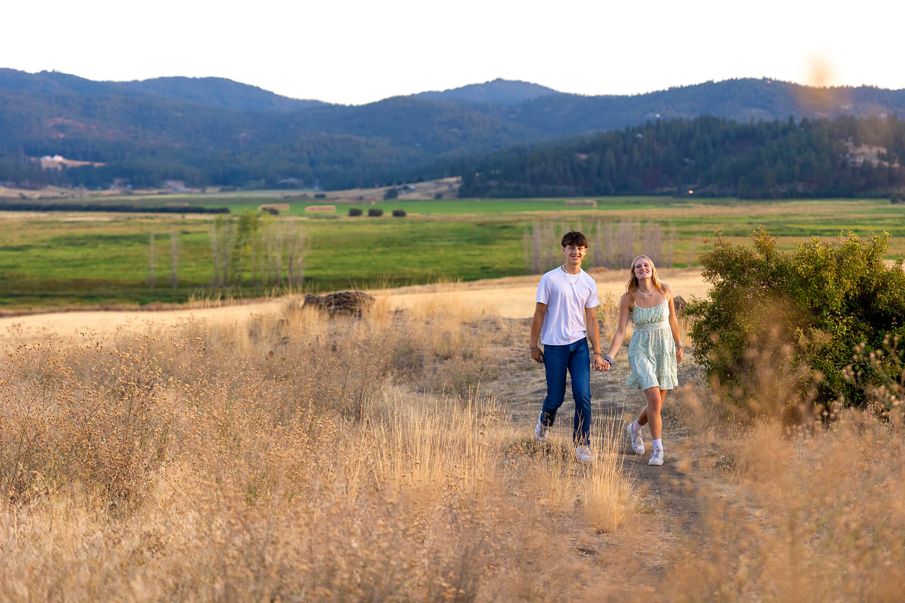 A blonde high school senior walking with her boyfriend in a grassy mountain setting wearing a short dress, white shoes and a watch boutiques in spokane