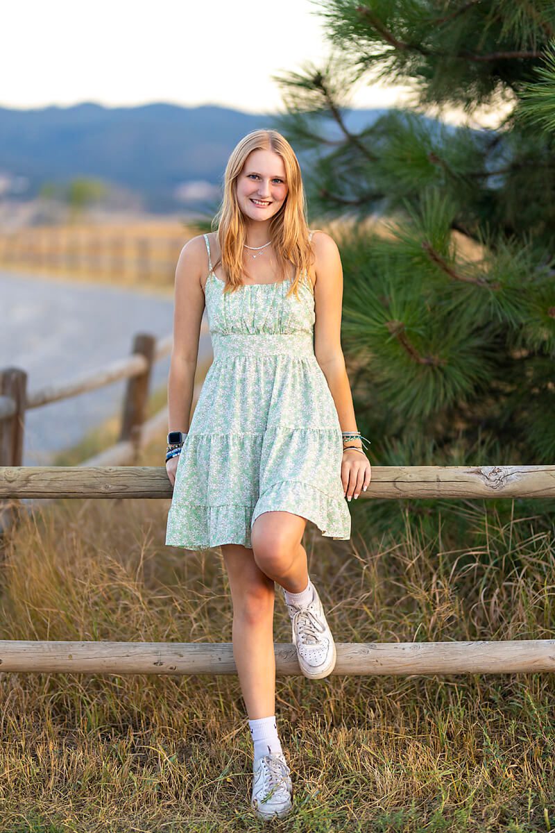 A high school senior leans along a fence line wearing a green dress, white shoes and accessories.