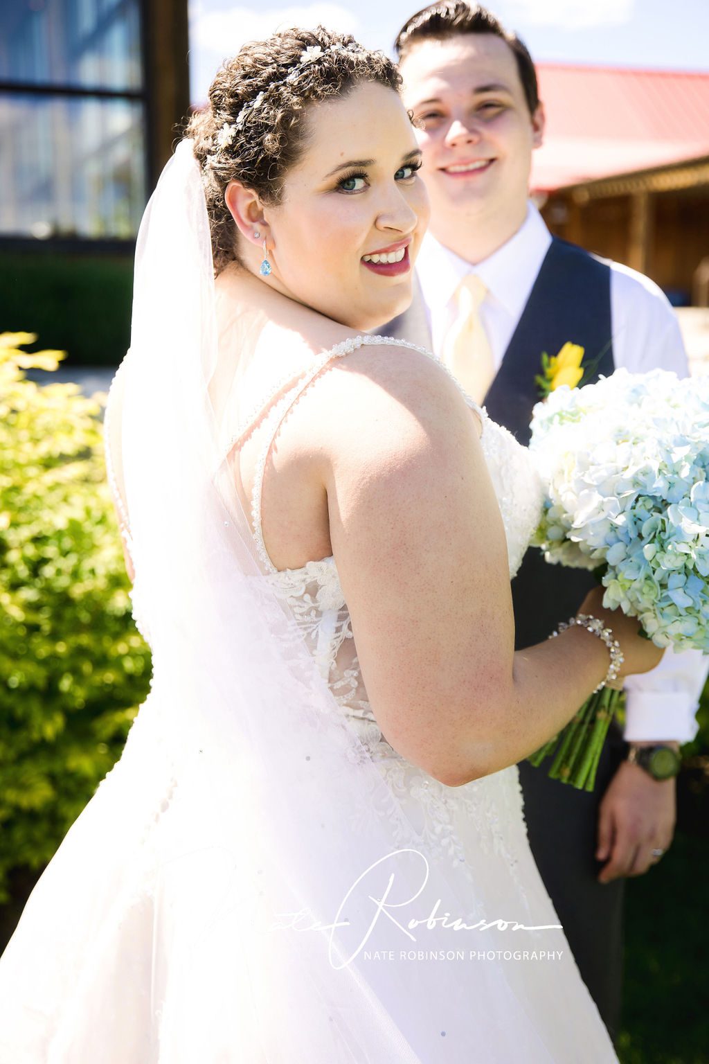 Bride looks over her shoulder wearing a long veil and white lace dress with blue bouquet red barn farm wedding venue