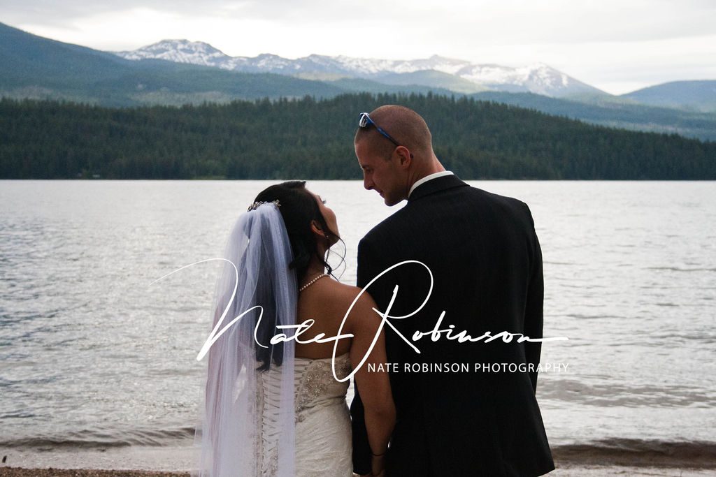 newlyweds stand on the edge of a large lake with a snowy mountain view zephyr lodge wedding