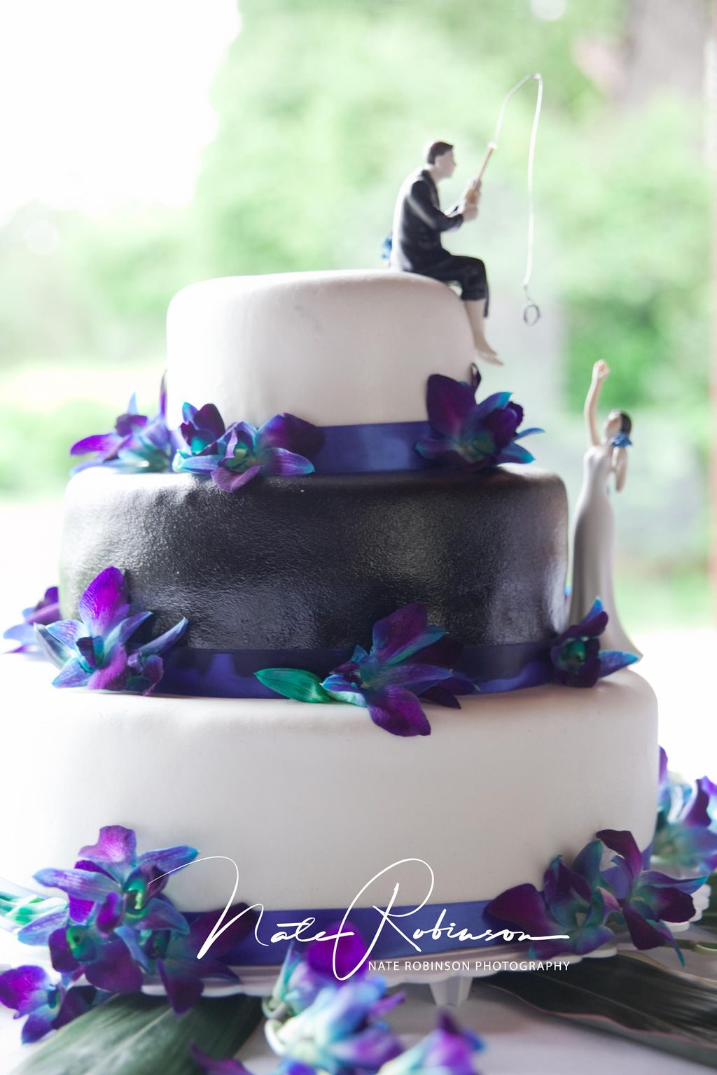 A three tier wedding cake decorated with purple flowers and figurings of groom fishing for bride with a ring zephyr lodge wedding