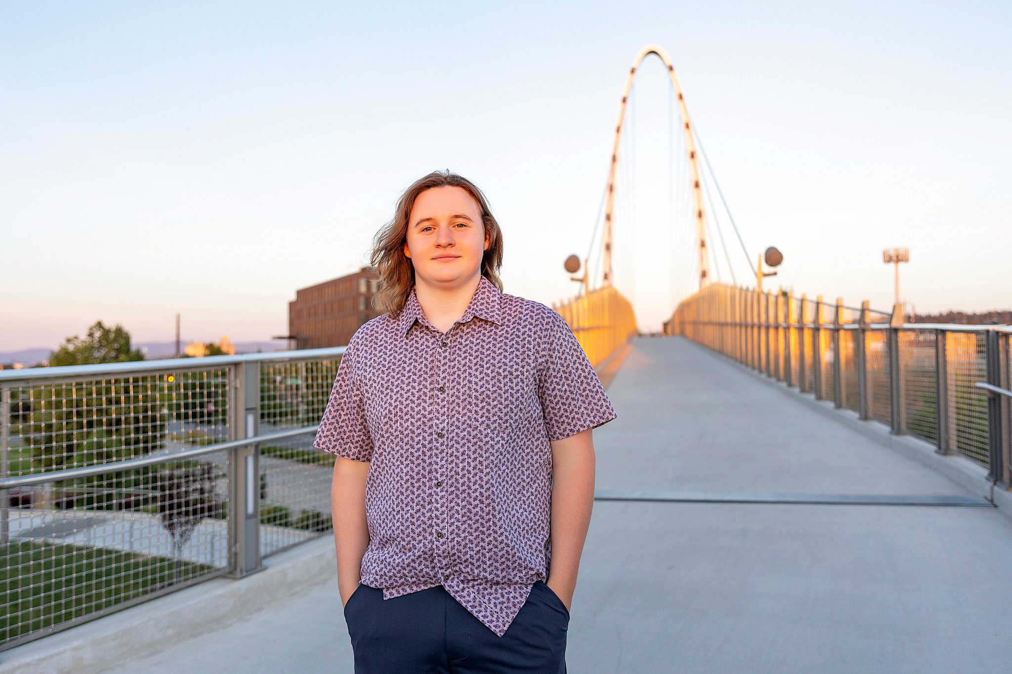 A high school senior in a button-down shirt walks down a pedestrian bridge at sunset