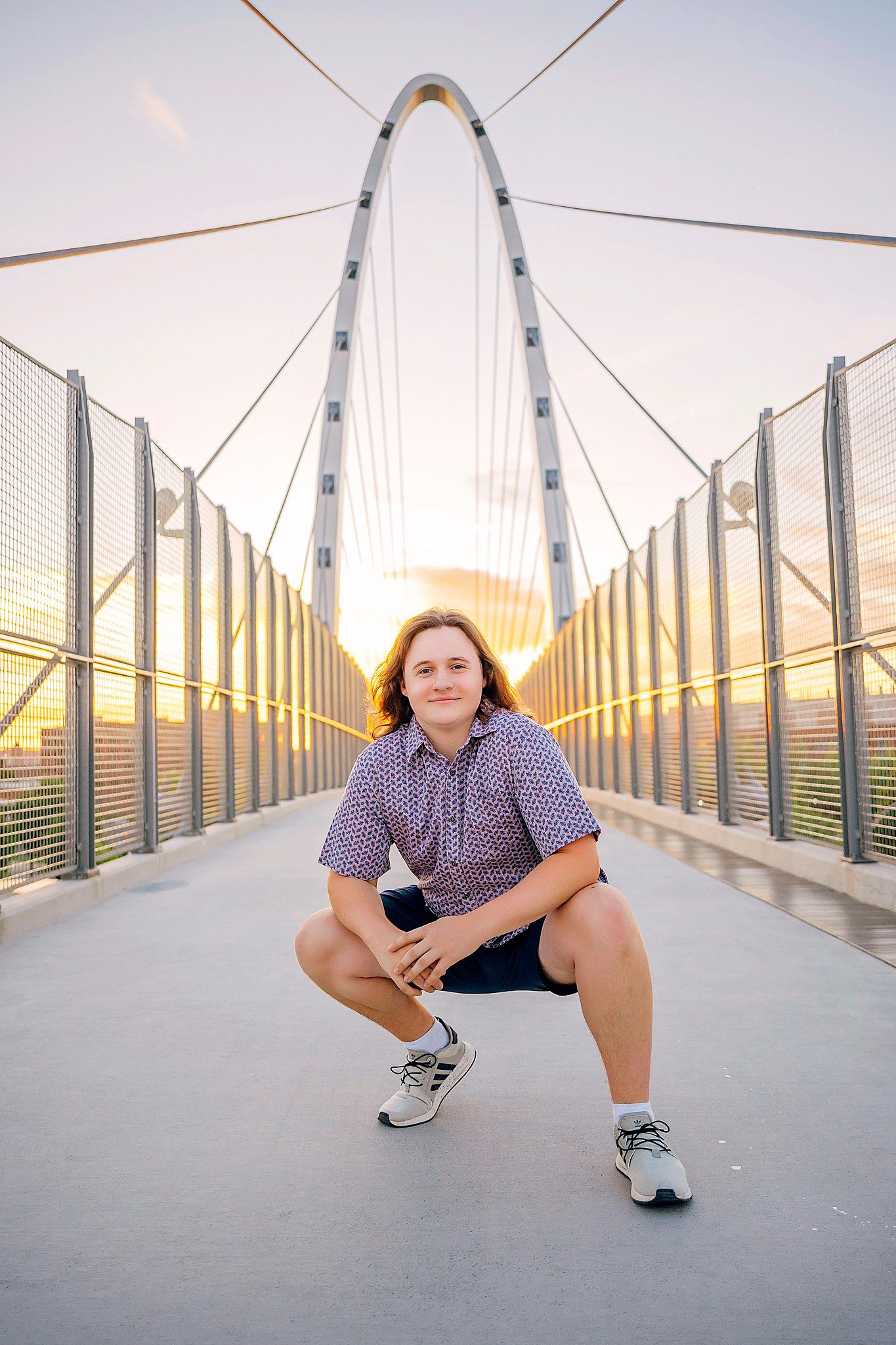 A high school senior with long hair and a button-down shirt and shorts squats in the center of a pedestrian suspension bridge colleges in spokane