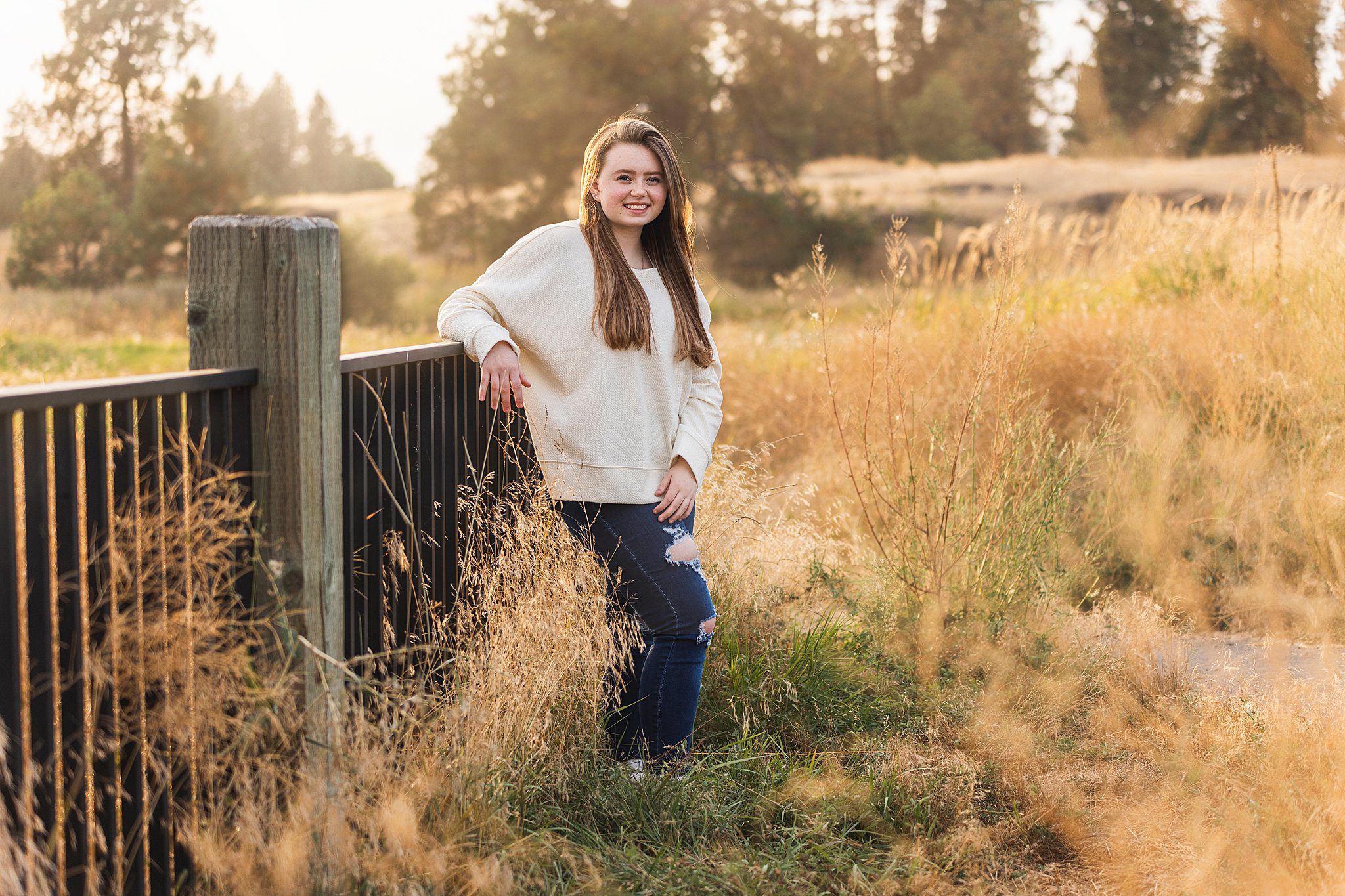 A high school senior stands against a metal fence with wooden posts in a golden field at sunset