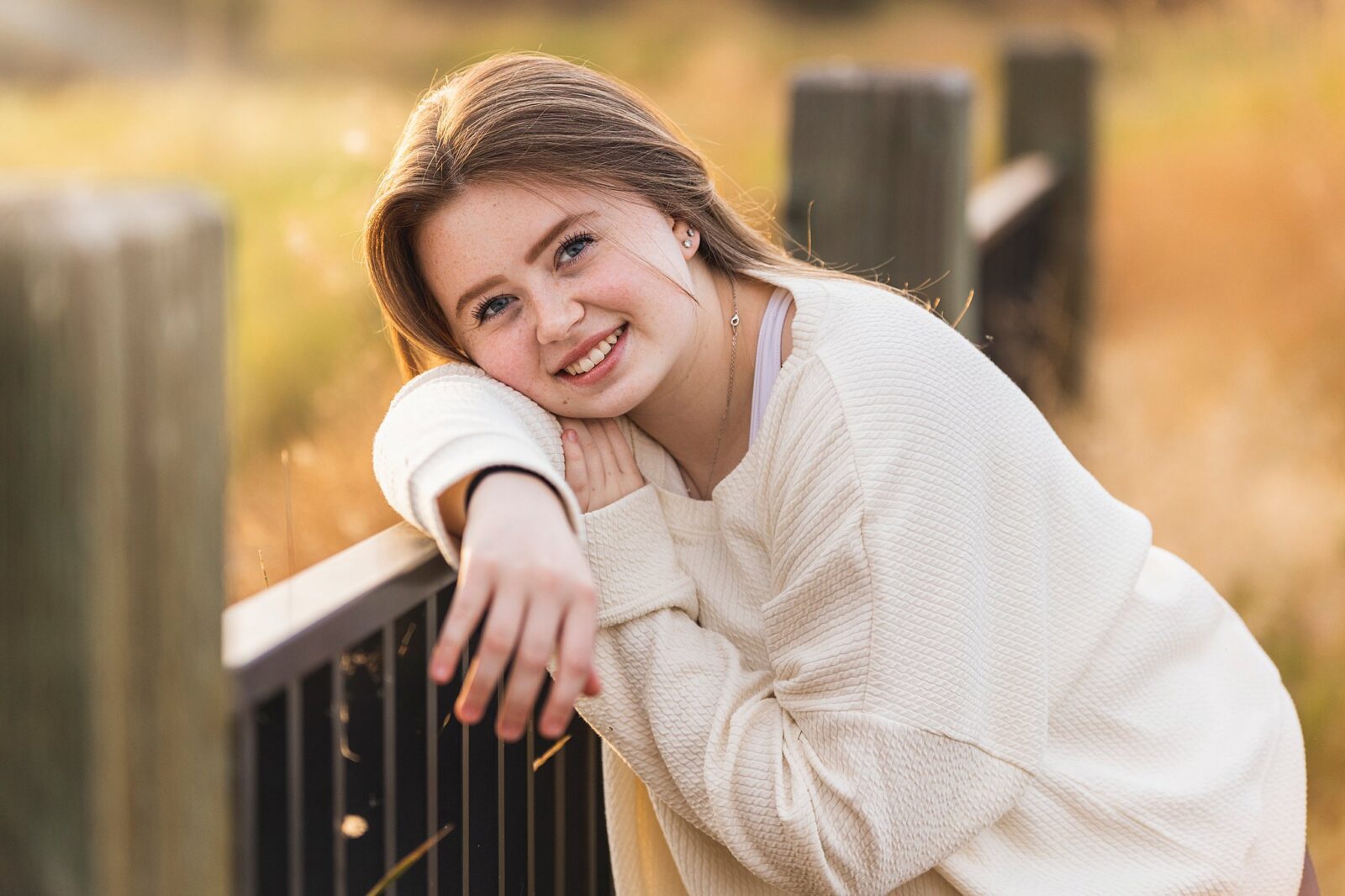 A high school senior leans against a modern fence in a golden field wearing a white sweater community colleges in spokane washington