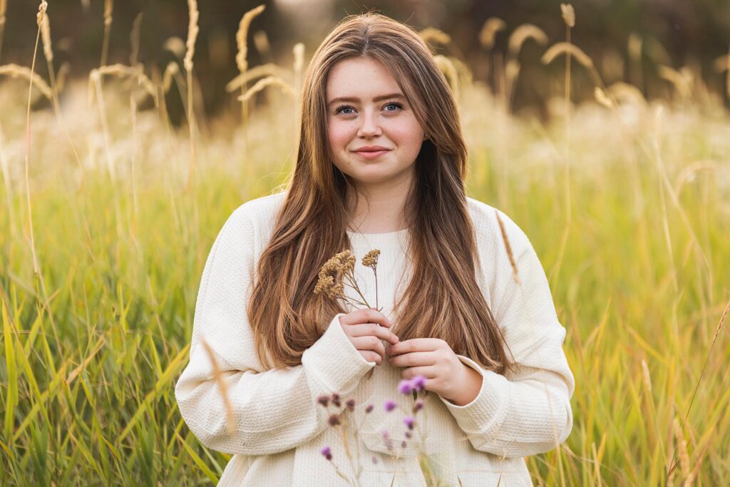 A high school senior sits in a field of tall grass holding a bundle small wildflowers