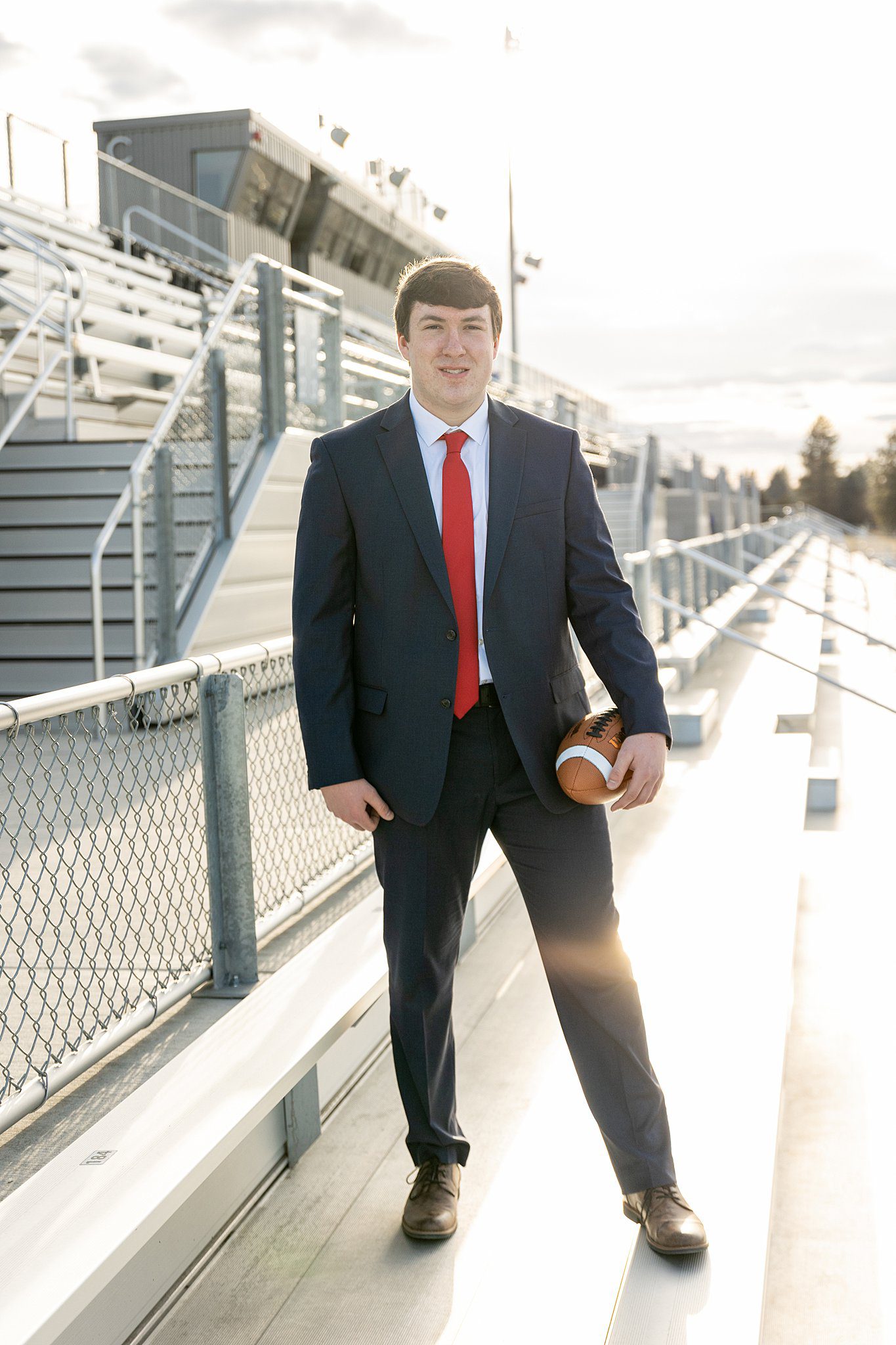 A high school senior stands in a full suit and red tie with a football in the stands of the stadium prom dress shops spokane