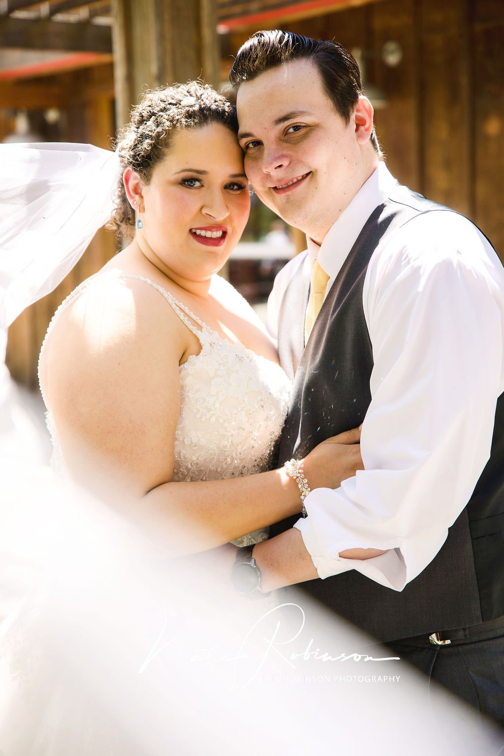 A long white veil flies in front of a newlywed couple wearing a white lace wedding dress and a black vest and yellow tie rustic river wedding venue