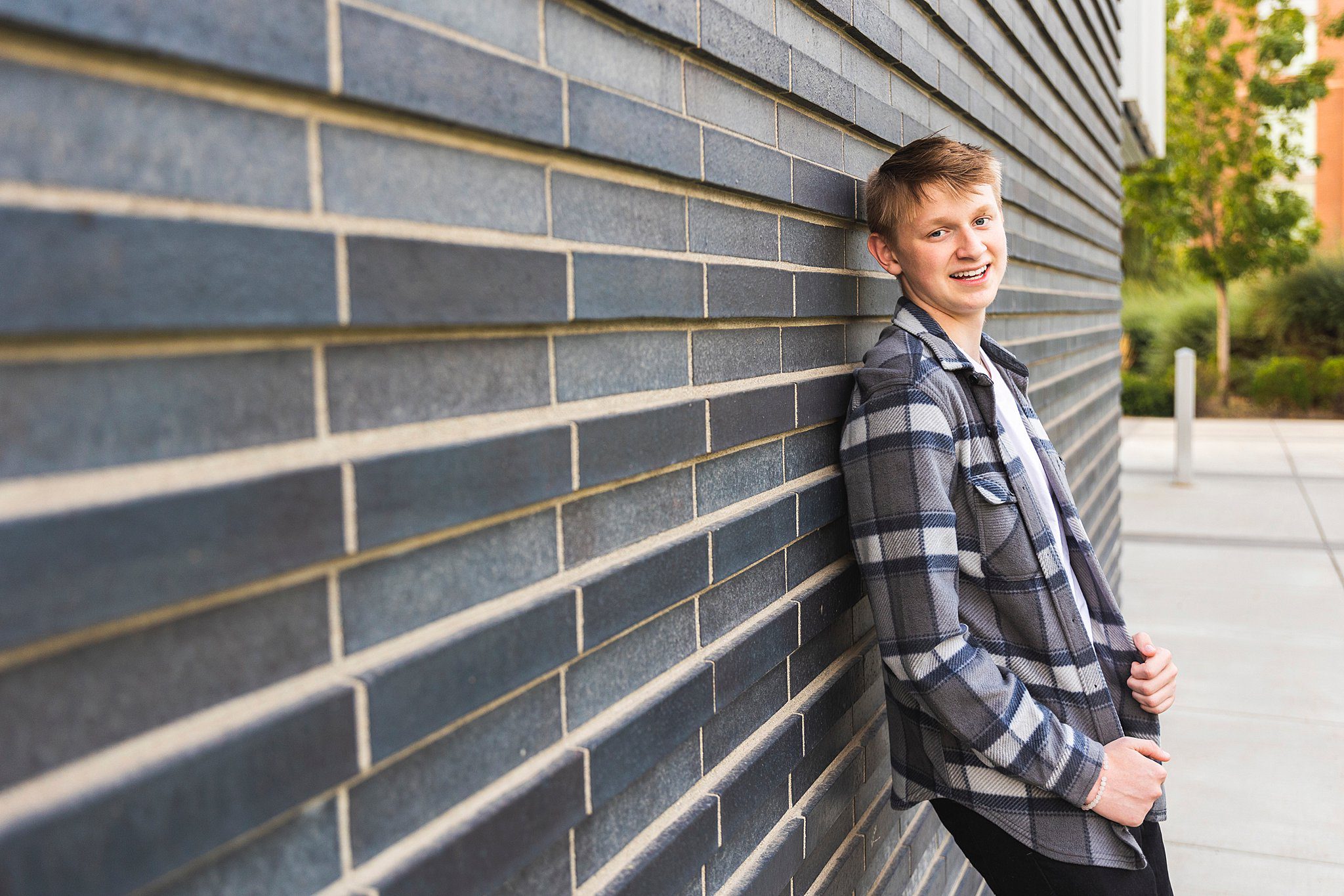 A high school senior boy leans against a dark brick wall wearing a blue flannel shirt scc running start