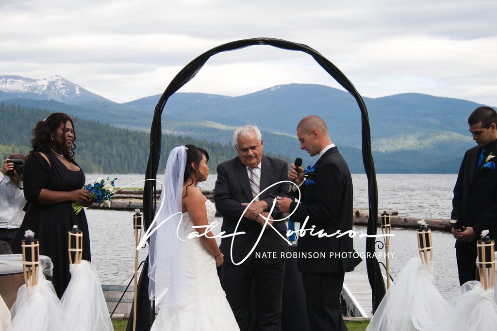 Bride and groom stand under an arbor on a dock exchanging vows with a mountain and waterfront background