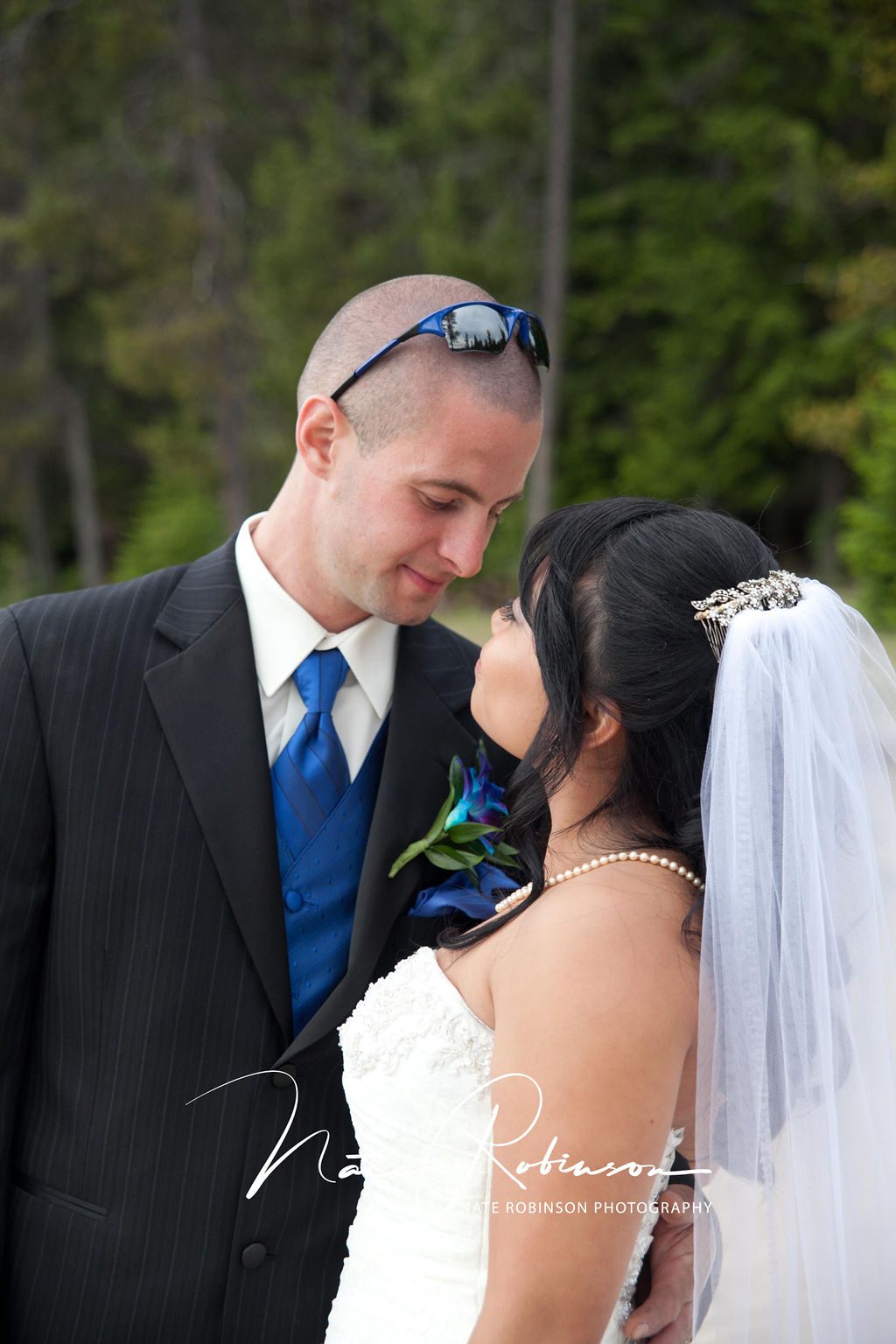 A groom wearing a blue vest and tie matching his boutonniere leans in for a kiss with his bride in a white lace dress and veil at a schweitzer wedding