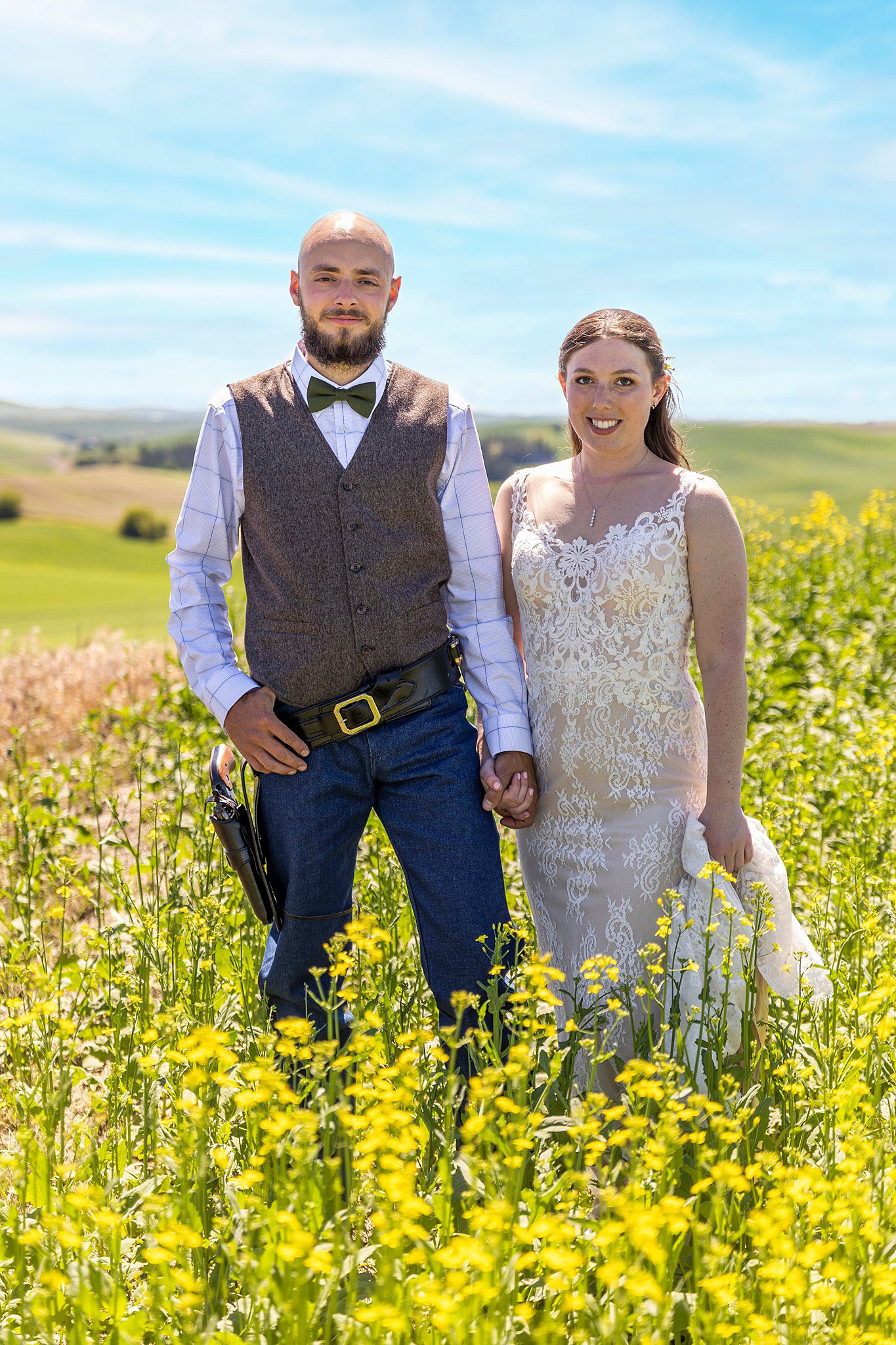 Newlyweds stand in a field of wildflowers holding hands wearing a lace wedding gown and a brown vest with a gun holster silverbell wedding
